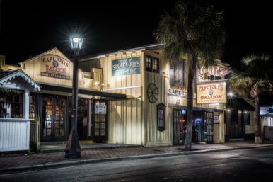 Historic wooden saloon with vintage 1930s signage, warm interior lights, palm trees and a glowing streetlamp on a quiet tropical night.