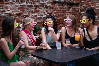 Five friends wearing colorful heart-shaped sunglasses, laughing and holding drinks on an urban brick-walled outdoor patio — playful bachelorette-style celebration.
