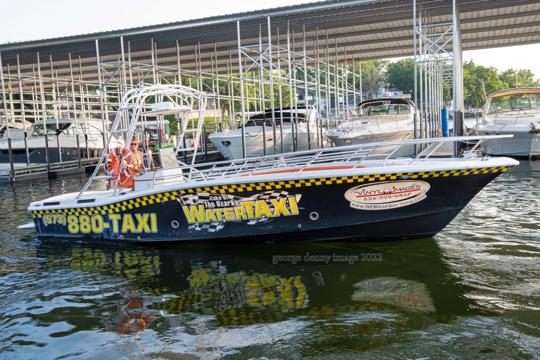 Black-and-yellow water taxi docked under a covered marina at Lake of the Ozarks, colorful signage and a life ring visible, reflected in rippling water