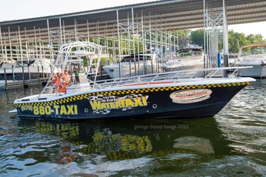 Black-and-yellow water taxi docked under a covered marina at Lake of the Ozarks, colorful signage and a life ring visible, reflected in rippling water