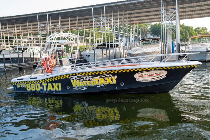 Black-and-yellow water taxi docked under a covered marina at Lake of the Ozarks, colorful signage and a life ring visible, reflected in rippling water