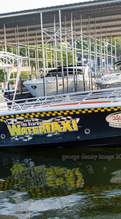 Black-and-yellow water taxi docked under a covered marina at Lake of the Ozarks, colorful signage and a life ring visible, reflected in rippling water