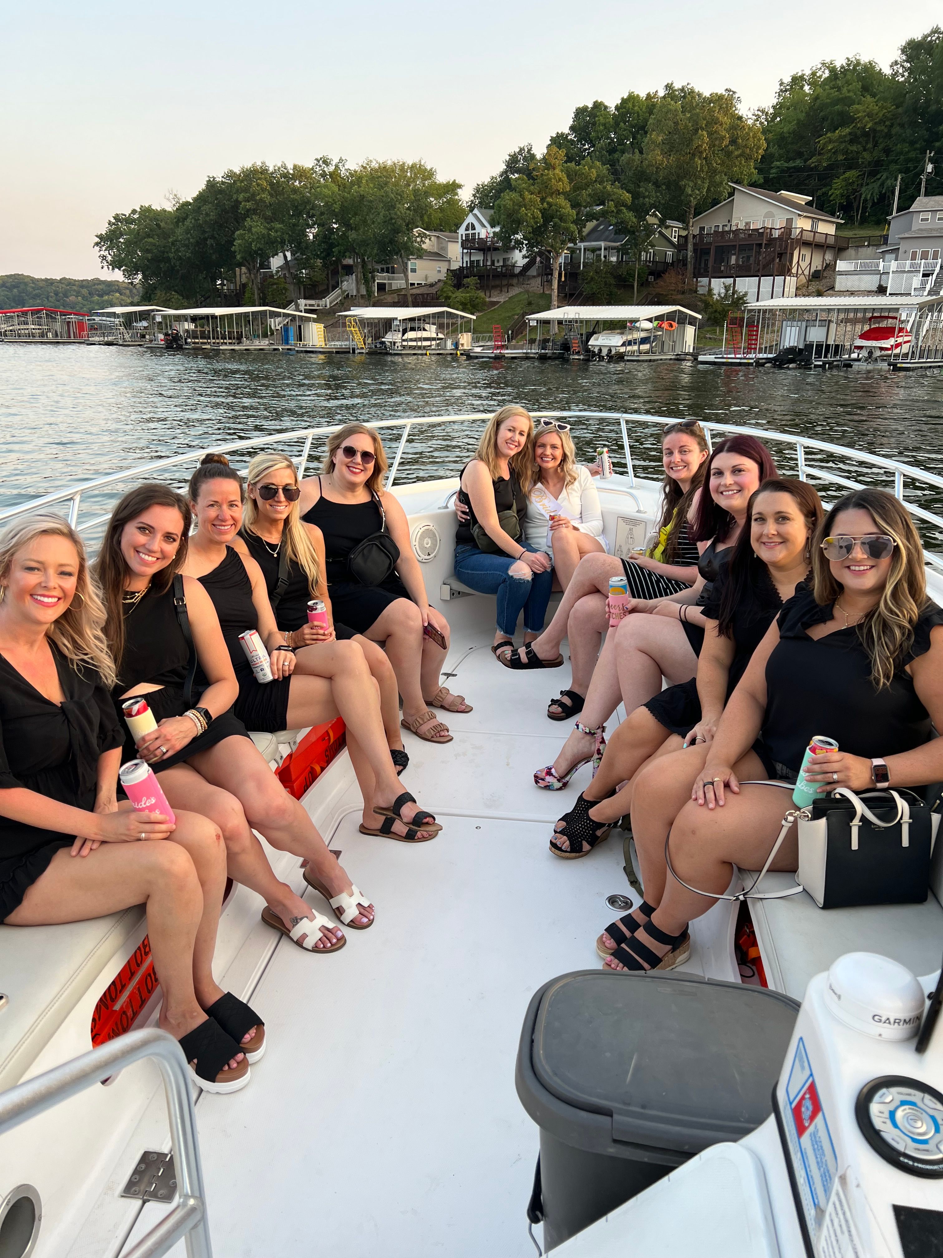Smiling group of women on a white motorboat enjoying drinks on a summer lake cruise at sunset, with lakefront houses and boat docks in the background.