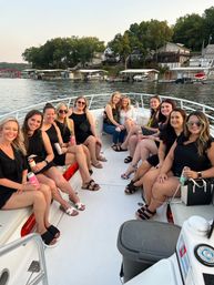 Smiling group of women on a white motorboat enjoying drinks on a summer lake cruise at sunset, with lakefront houses and boat docks in the background.