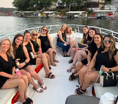 Smiling group of women on a white motorboat enjoying drinks on a summer lake cruise at sunset, with lakefront houses and boat docks in the background.