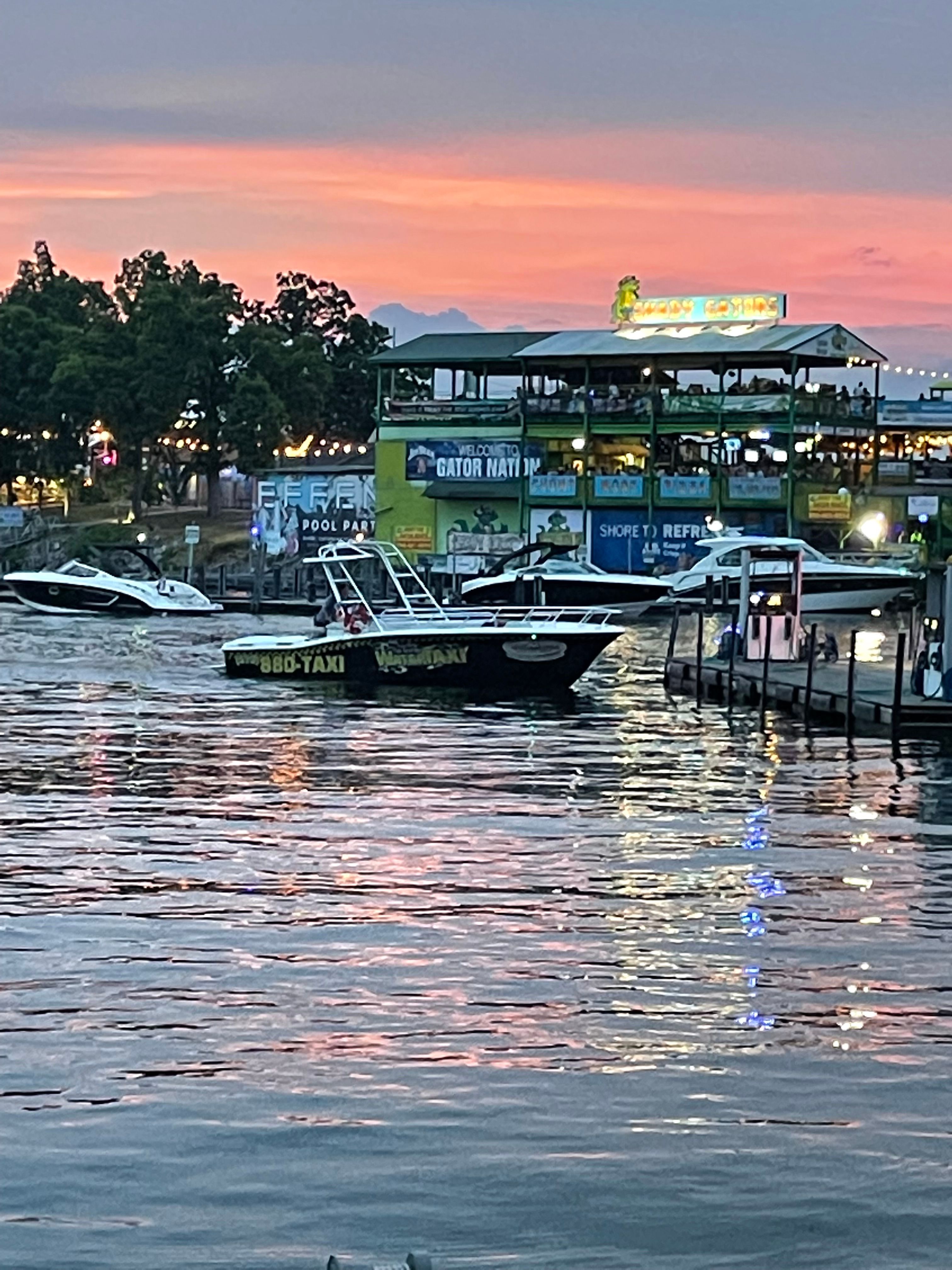 Evening waterfront marina at sunset with docked boats and a black water-taxi near a lit two-story riverside restaurant, pink-orange sky and shimmering reflections on rippling water.