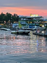 Evening waterfront marina at sunset with docked boats and a black water-taxi near a lit two-story riverside restaurant, pink-orange sky and shimmering reflections on rippling water.