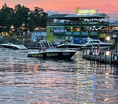 Evening waterfront marina at sunset with docked boats and a black water-taxi near a lit two-story riverside restaurant, pink-orange sky and shimmering reflections on rippling water.