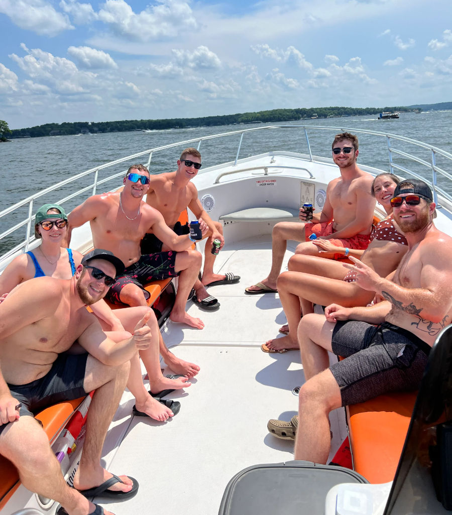 Group of friends in swimsuits smiling and holding drinks on the bow of a powerboat cruising a sunny lake with tree-lined shoreline and blue sky