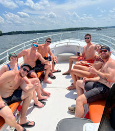 Group of friends in swimsuits smiling and holding drinks on the bow of a powerboat cruising a sunny lake with tree-lined shoreline and blue sky