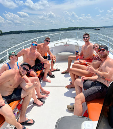 Group of friends in swimsuits smiling and holding drinks on the bow of a powerboat cruising a sunny lake with tree-lined shoreline and blue sky
