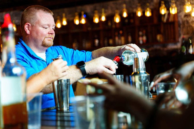 Bartender pouring clear liquor into a jigger behind a wooden bar, surrounded by cocktail shakers, bottles, and warm Edison-bulb lighting — lively cocktail bar scene.