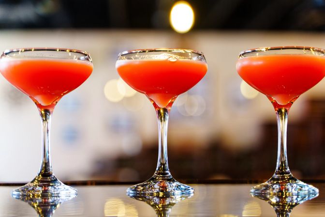 Three coral-orange cocktails in coupe glasses lined up on a glossy bar counter, reflected below with warm bokeh lights in the background.