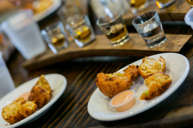Close-up of Southern-style fried corn fritters on white plates with a creamy dipping sauce, set on a dark wooden table with a whiskey tasting flight blurred in the background.