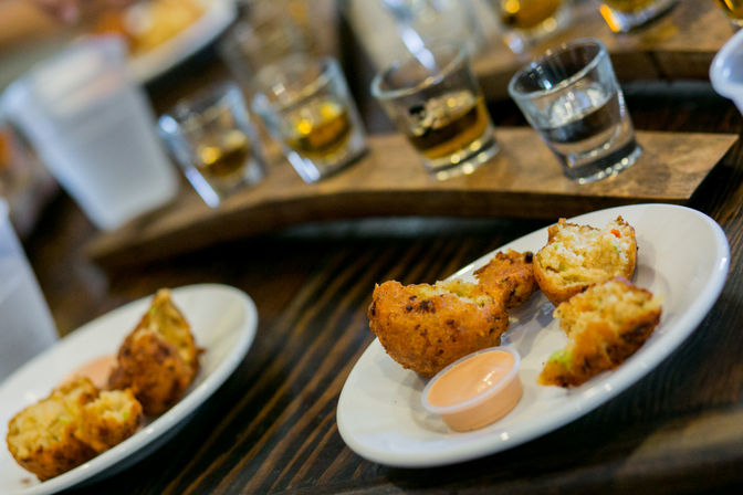 Close-up of Southern-style fried corn fritters on white plates with a creamy dipping sauce, set on a dark wooden table with a whiskey tasting flight blurred in the background.