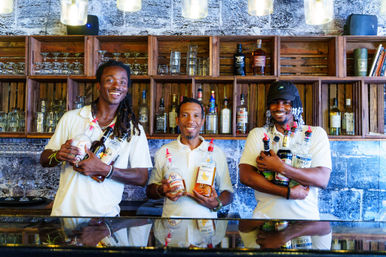 Three smiling bartenders behind a rustic bar holding assorted rum and liquor bottles in front of wooden shelves stacked with glassware and spirits, colorful island-style vibe.