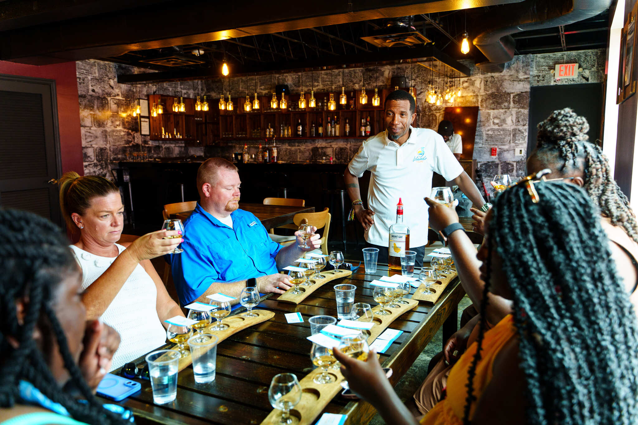 Group of adults enjoying a spirits tasting flight at a cozy craft distillery tasting room — wooden flight boards and tasting glasses on a long table, bartender explaining samples under warm Edison-bulb lighting and exposed brick bar.