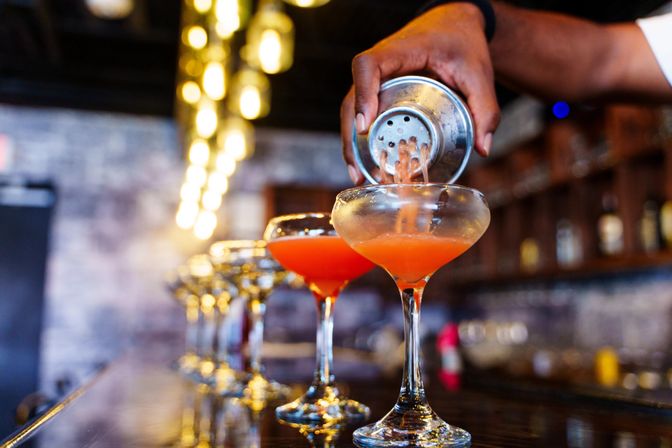 Bartender's hand pouring an orange-red craft cocktail from a metal shaker into a row of coupe glasses on a glossy bar counter under warm string lights in a cozy urban bar.