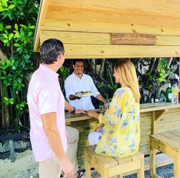 Smiling bartender serves a plate to a couple at a wooden outdoor beach bar surrounded by lush tropical foliage and bottles on the counter.