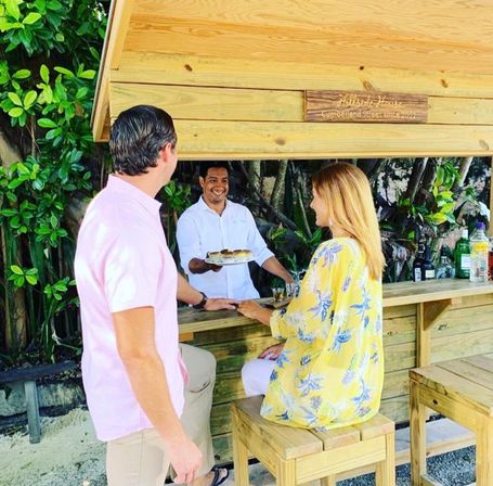 Smiling bartender serves a plate to a couple at a wooden outdoor beach bar surrounded by lush tropical foliage and bottles on the counter.