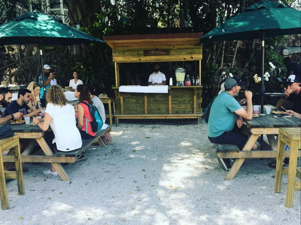 Sun-dappled outdoor café with a wooden kiosk and green umbrellas, casual diners at picnic tables on a gravel patio surrounded by tropical foliage and a server at a juice station.