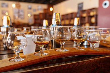 Whiskey tasting flight — row of snifter glasses with amber spirits on a wooden paddle in a warm-lit distillery tasting room.