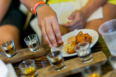 Hand reaching for a crispy golden hush puppy on a white plate with dipping sauce, surrounded by a whiskey shot flight on wooden paddles at a casual tasting.