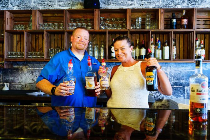Two smiling bartenders behind a rustic bar counter holding liquor bottles, wooden shelves of glassware and bottles, exposed stone wall and glossy countertop reflection.
