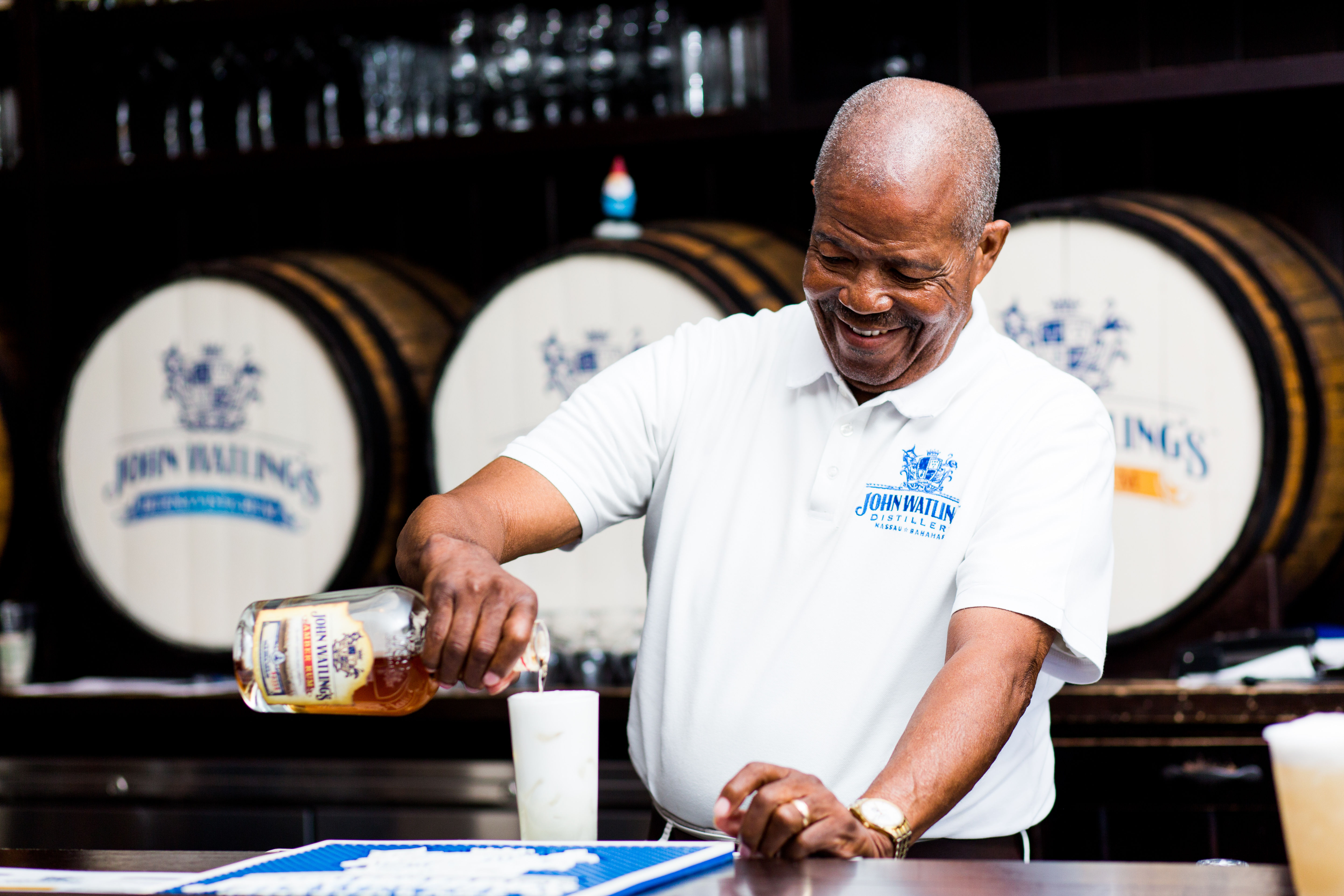 Smiling bartender pouring rum into a tall glass at a distillery-style bar, with wooden aging barrels stacked in the background.