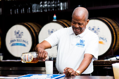 Smiling bartender pouring rum into a tall glass at a distillery-style bar, with wooden aging barrels stacked in the background.