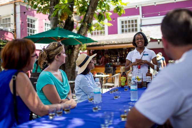 Outdoor rum-tasting tour in a sunny, colorful courtyard — a smiling guide presents bottles on a blue tablecloth while a small group samples shots under green umbrellas.