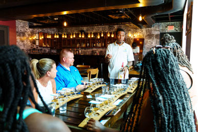 Guide-led spirits tasting in a cozy urban tasting room — host pouring from a bottle at a long wooden table set with wooden flight boards and small tasting glasses, warm industrial pendant lights and exposed brick walls.