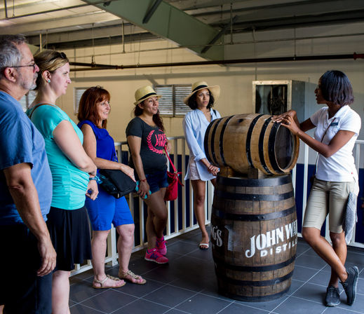 Small tour group listening to a guide beside stacked wooden barrels inside an indoor distillery warehouse, visitors in summer clothes and sun hats.