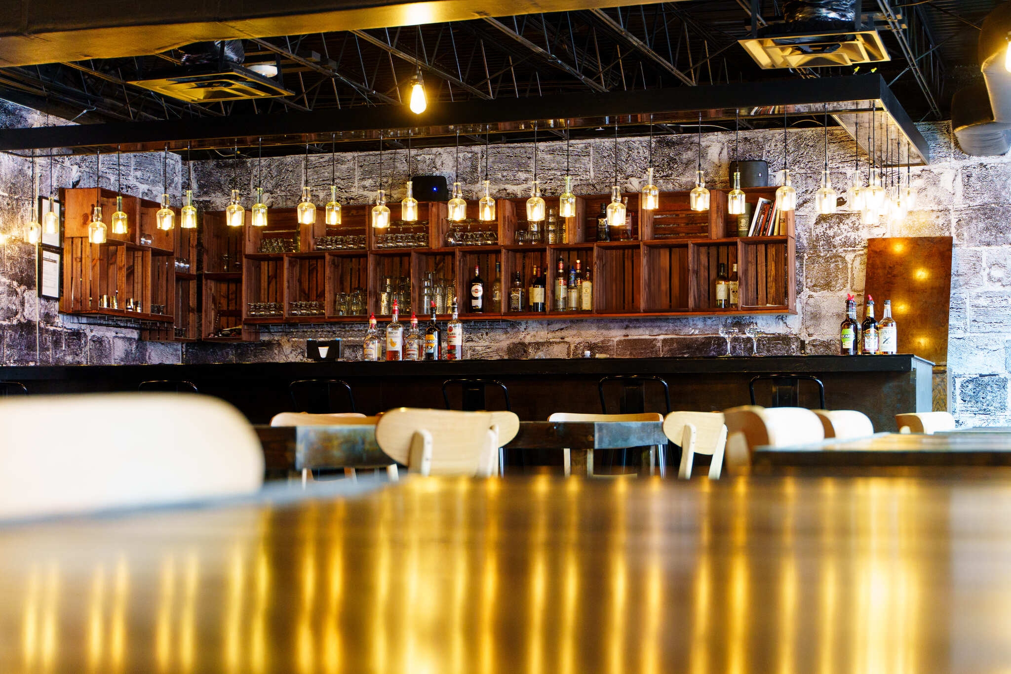 Cozy industrial bar interior with warm Edison pendant lights, wooden shelving stocked with bottles, a textured stone wall backdrop, bar counter and empty chairs with golden reflections on a polished table.