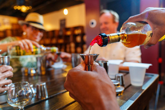 Close-up of amber whiskey being poured into a metal jigger at a lively tasting bar, with blurred patrons, tasting glasses, and a wooden counter in the background.