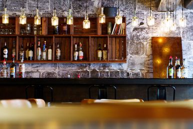 Cozy industrial-style bar interior with exposed brick, wooden shelves of liquor bottles, amber pendant lights, and coupe glasses lined up on a dark counter.