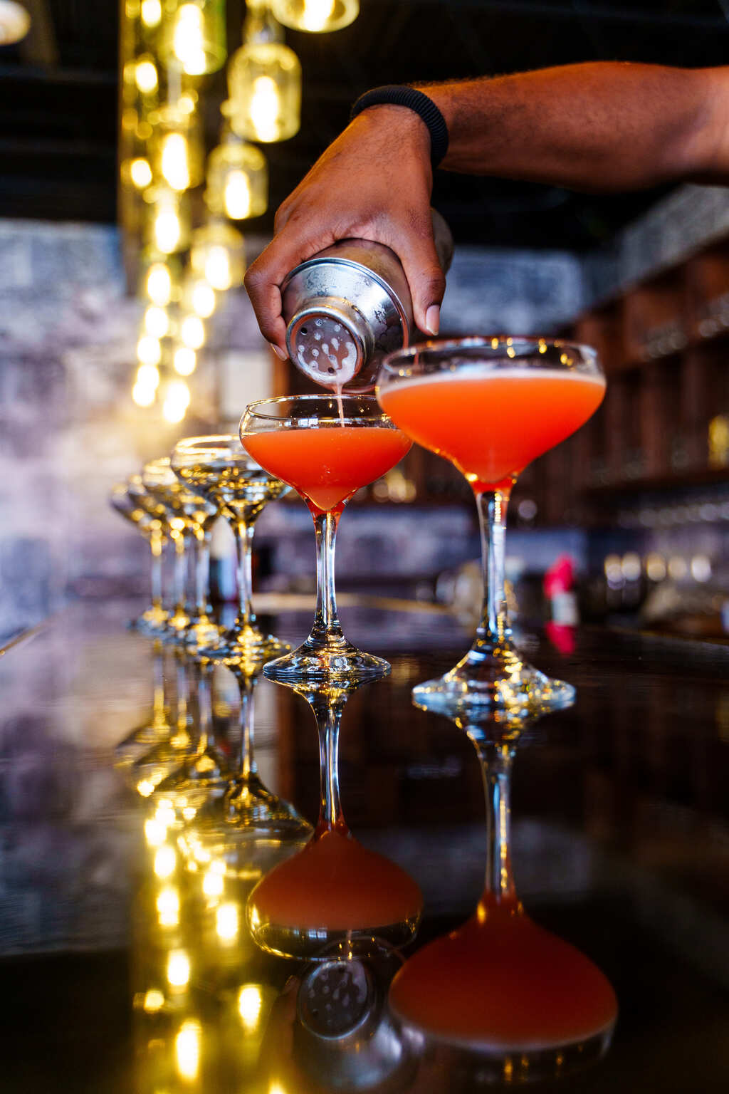 Hand pouring vibrant orange craft cocktails from a metal shaker into coupe glasses on a glossy bar countertop, with a row of glasses and warm bokeh lights in the background.