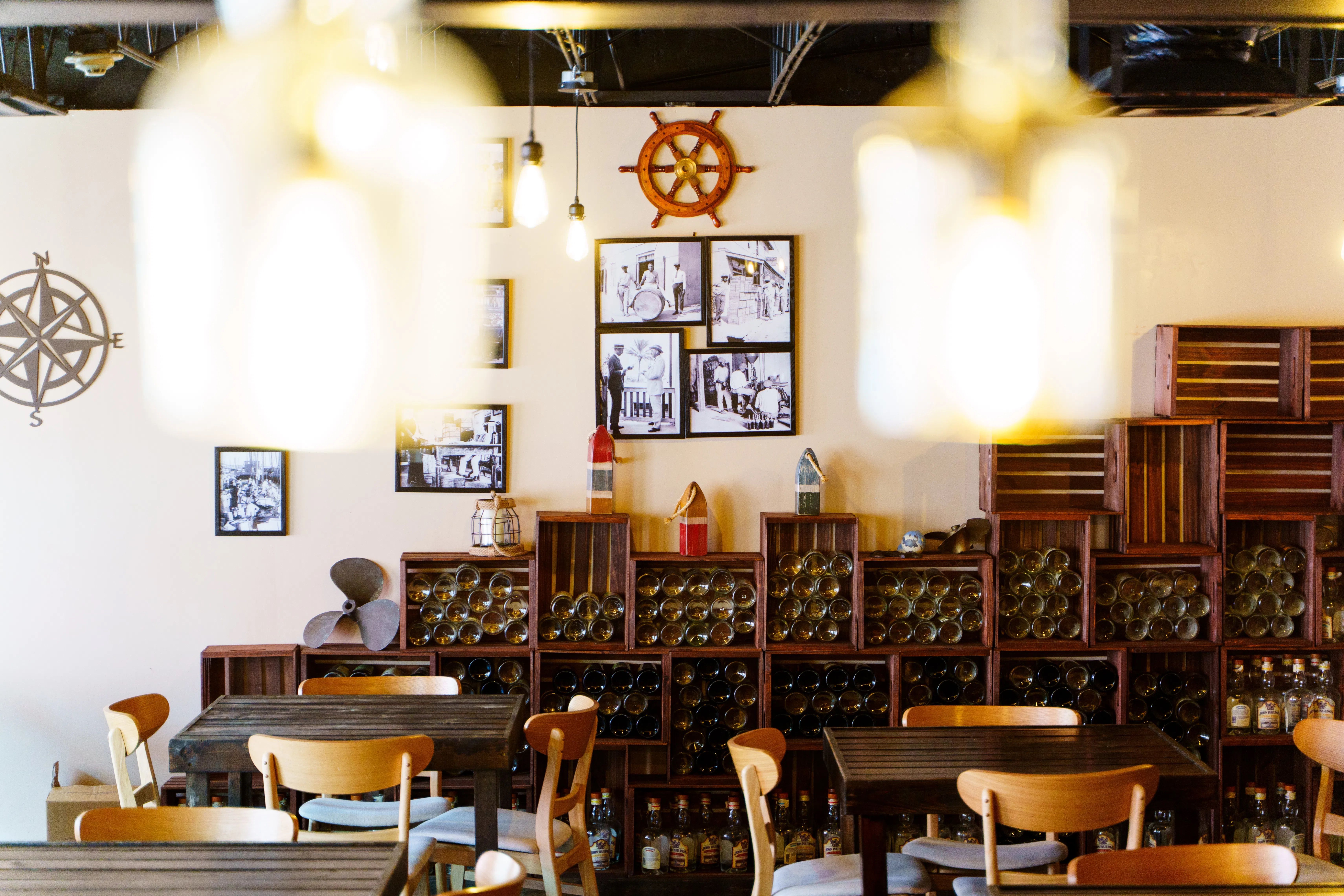 Cozy nautical-themed wine bar interior with rustic wooden tables and chairs, stacked wine bottles in crate shelving, ship wheel and compass wall decor, warm pendant lights and vintage black-and-white photos.