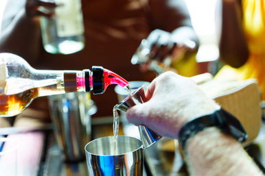 Close-up of hands pouring golden liquor from a bottle into a metal jigger over a stainless cocktail shaker at a busy bar