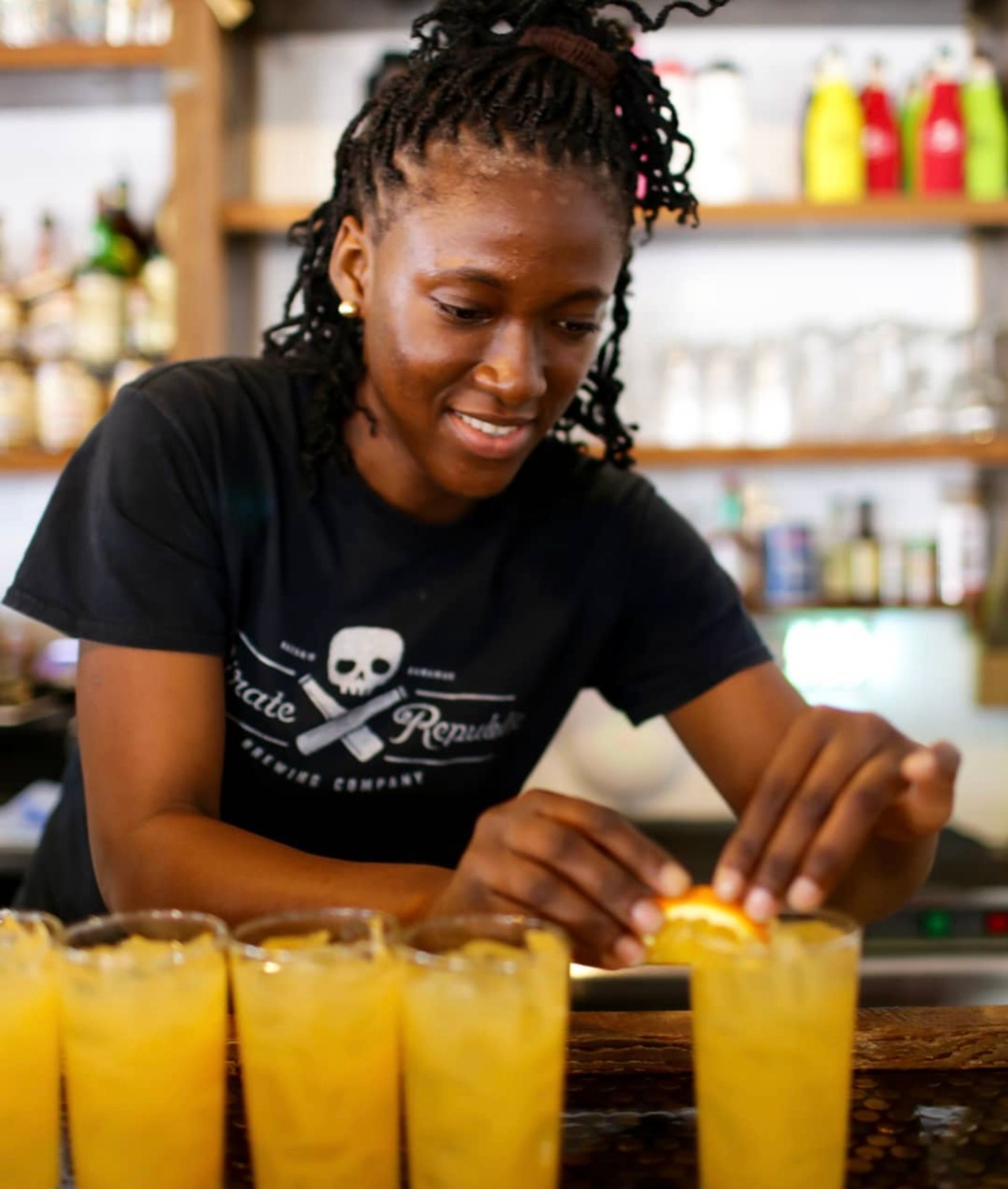 Smiling bartender garnishing a row of orange cocktails with citrus slices on a wooden bar counter, bottles and glassware on shelves in the background