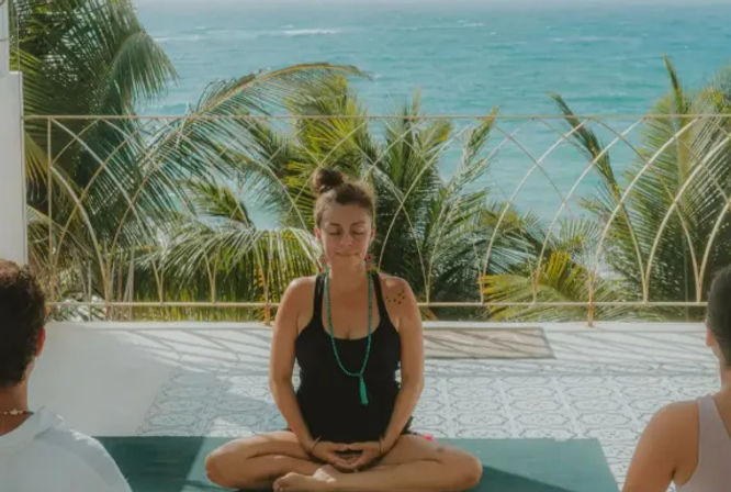 Oceanfront yoga: person meditating cross-legged on a tropical balcony with palm trees and turquoise sea in the background, classmates partially visible.