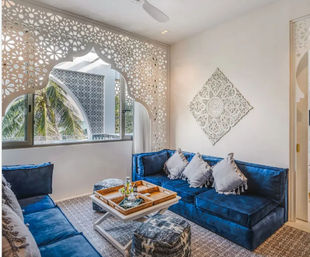 Sunlit coastal lounge with deep blue velvet sectional, tasseled throw pillows, white carved lattice screen and wall medallion, wooden tray on coffee table and palm trees visible through the window.