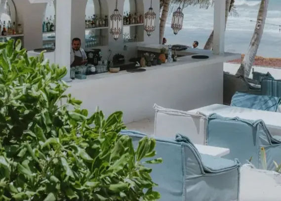 Beachfront outdoor bar with white counter and hanging lanterns, aqua cushioned lounge seating, leafy foreground and palm trees framing ocean waves in the background.