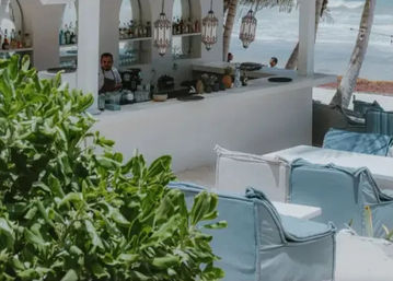 Beachfront outdoor bar with white counter and hanging lanterns, aqua cushioned lounge seating, leafy foreground and palm trees framing ocean waves in the background.