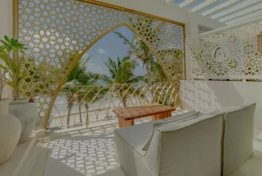 Sunny tropical beachfront terrace with cushioned lounge seating and a wooden table behind an ornate geometric lattice arch framing swaying palm trees and turquoise ocean.