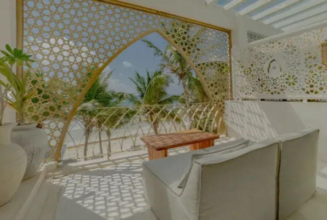 Sunny tropical beachfront terrace with cushioned lounge seating and a wooden table behind an ornate geometric lattice arch framing swaying palm trees and turquoise ocean.