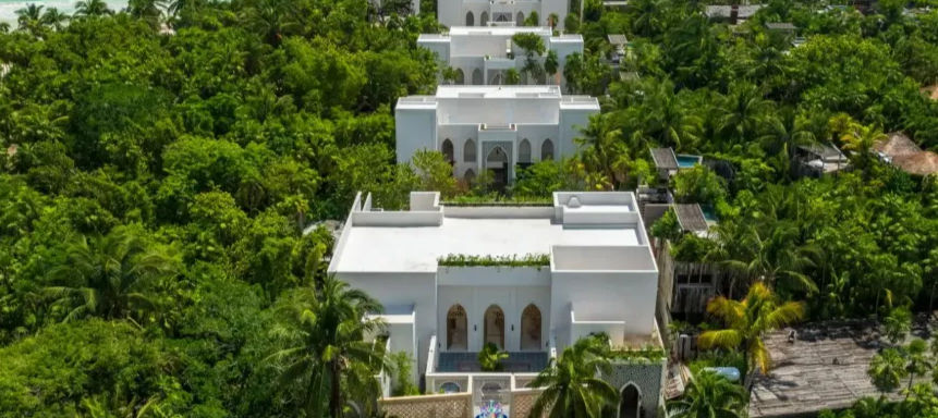 Drone-shot of pristine white villas tucked into a lush tropical palm forest, repeating courtyards and terraces amid dense green foliage.