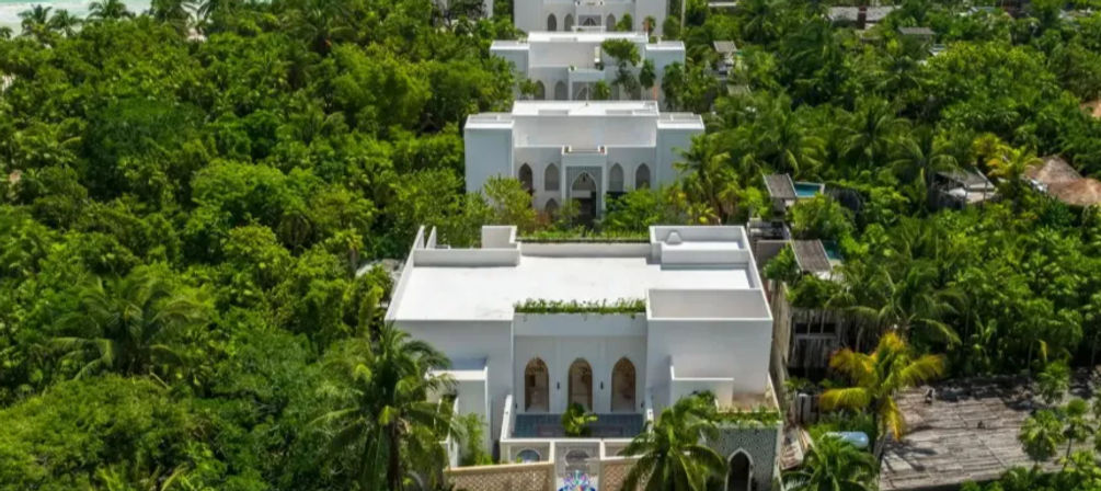 Drone-shot of pristine white villas tucked into a lush tropical palm forest, repeating courtyards and terraces amid dense green foliage.