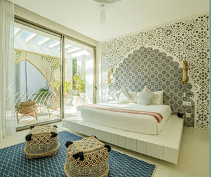 Sunlit boho-Moroccan bedroom with scalloped patterned tile headboard, low white platform bed, gold wall sconces, sliding glass doors to a small patio with rattan chairs, blue rug and woven poufs.