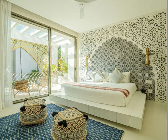 Sunlit boho-Moroccan bedroom with scalloped patterned tile headboard, low white platform bed, gold wall sconces, sliding glass doors to a small patio with rattan chairs, blue rug and woven poufs.
